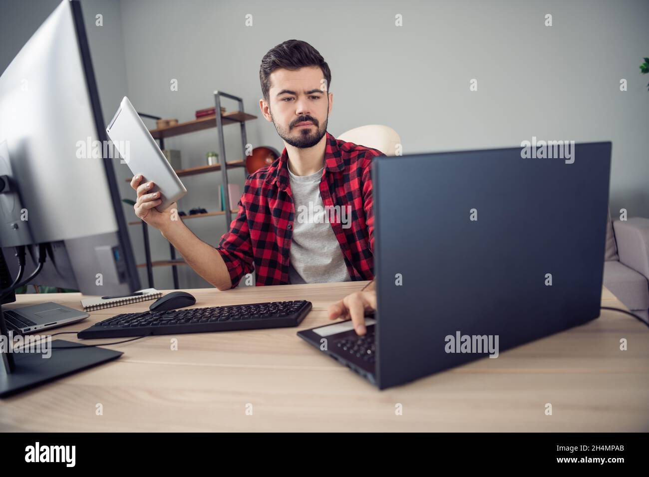 Photo of serious concentrated young man look laptop write hold tablet sit desk inside office indoors Stock Photo