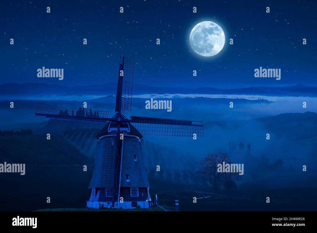 Night landscape with windmill on hill. Fog in valley and beautiful stars in sky Stock Photo