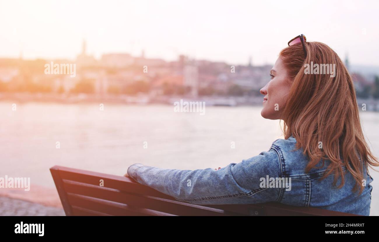Young balanced redhead Caucasian woman sitting on bench relaxing at riverside in sunset Stock Photo