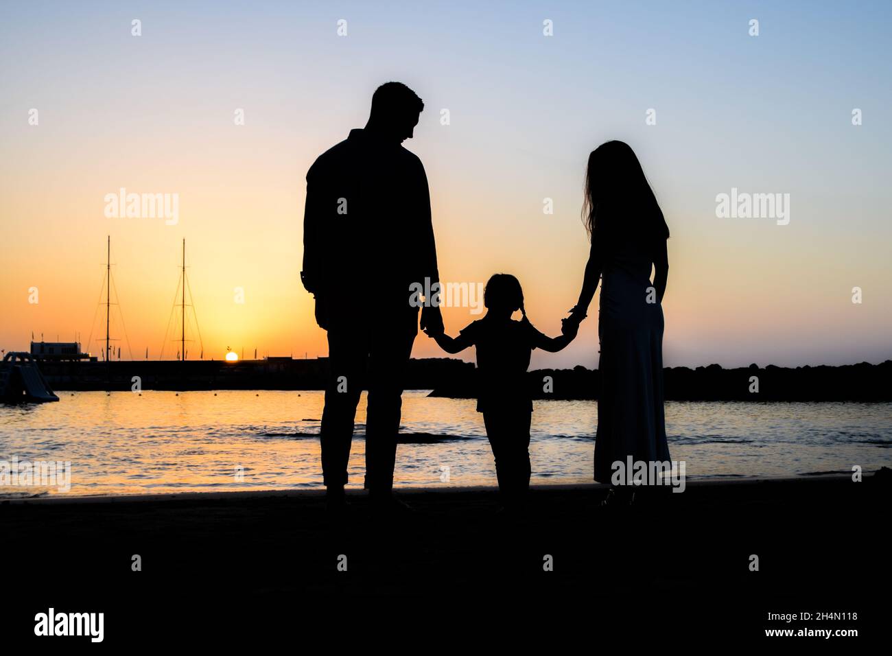 silhouette of a family at the beach holding hands during sunset. Father and mother look at their daughter. Romantic and passionate family moment Stock Photo