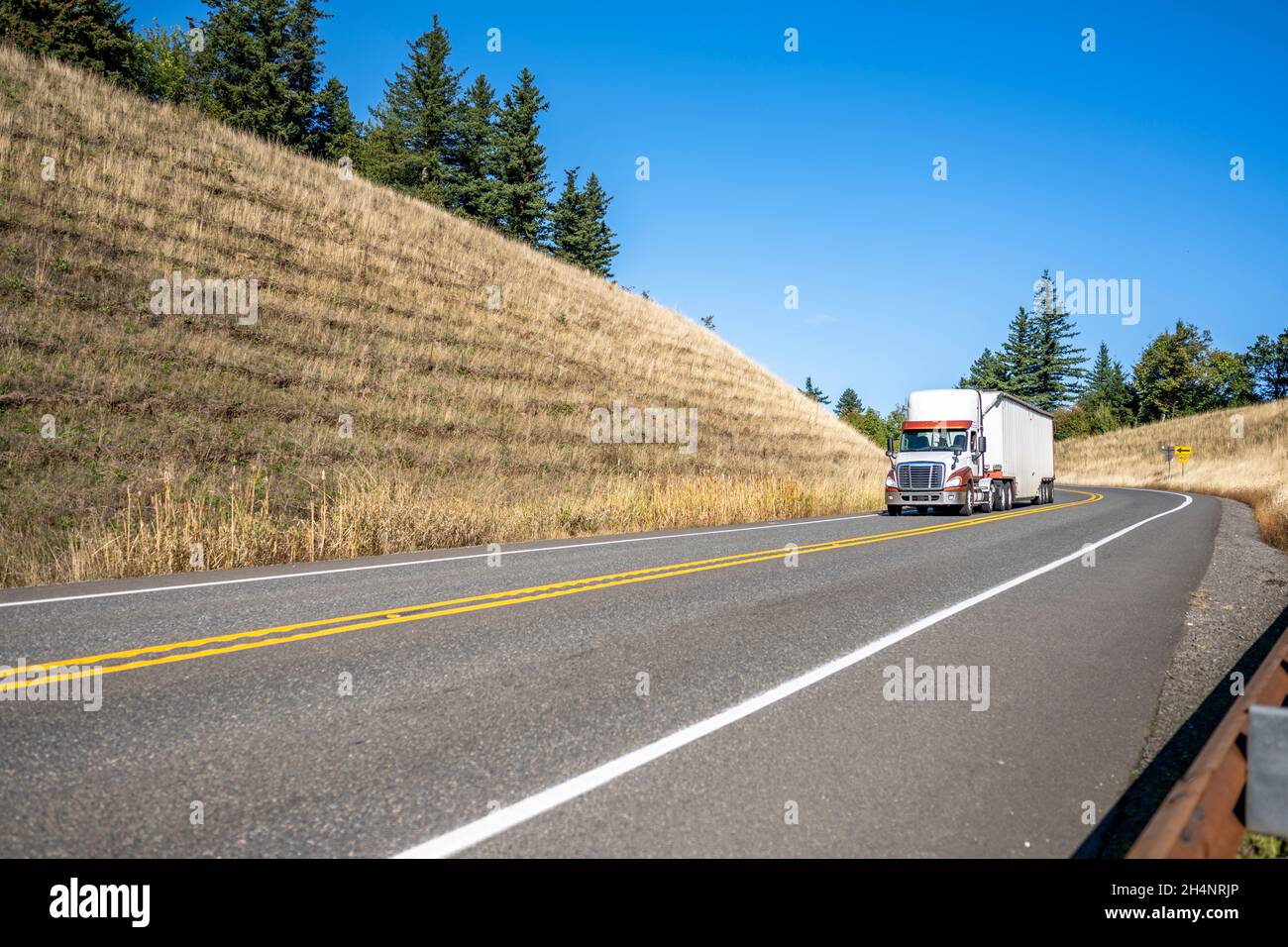 Big rig white semi truck with high cab transporting commercial cargo in covered bulk semi trailer running for delivery turning on the winding mountain Stock Photo