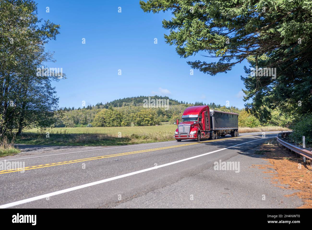Classic big rig red bonnet semi truck with high cab transporting commercial cargo in covered bulk semi trailer running for delivery on the winding nar Stock Photo