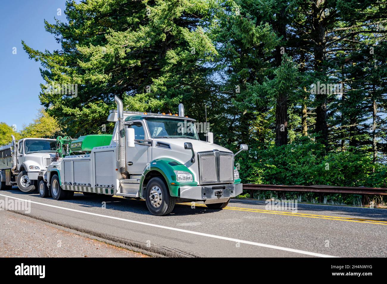 Green and white powerful road assistant big rig towing semi truck tow another broken semi truck transporting it to repair shop running on the road alo Stock Photo