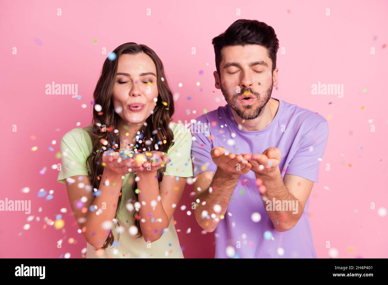 Portrait of attractive dreamy cheerful couple blowing decorative elements isolated over pink pastel color background Stock Photo