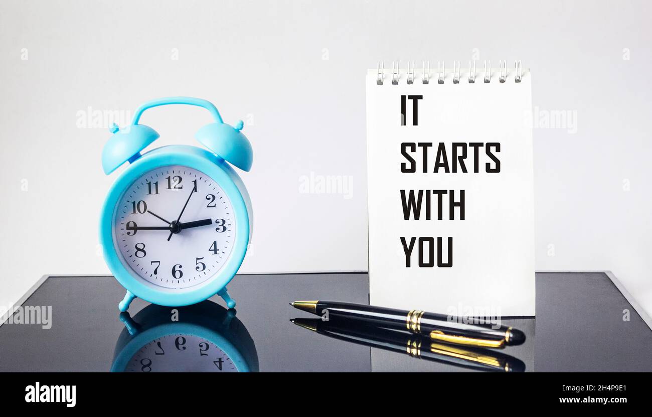 It all starts with you, text on a notebook, next to an alarm clock and a pen on a glass black background Stock Photo