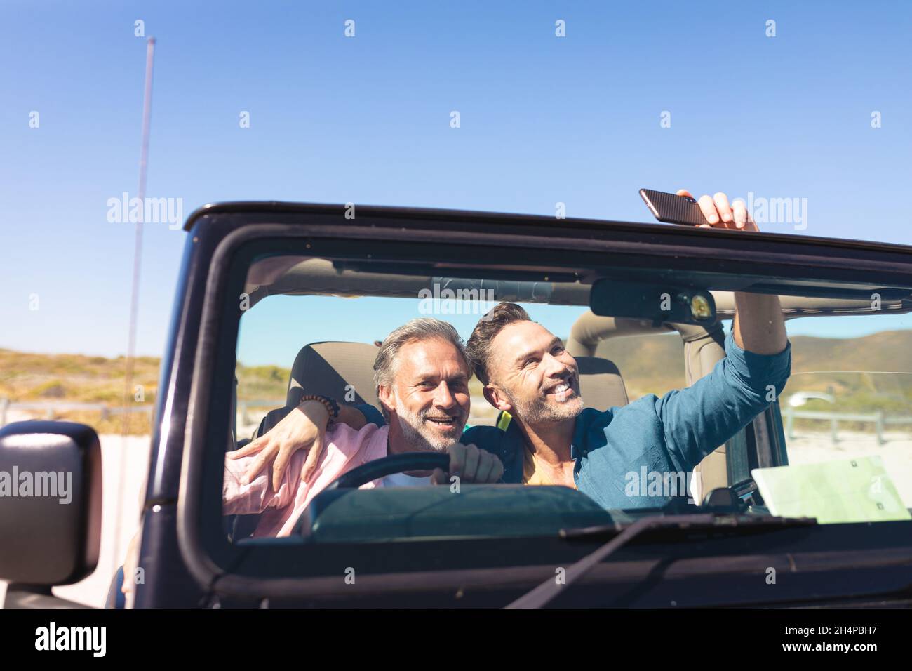 Smiling caucasian gay male couple taking selfies sitting in car at seaside Stock Photo