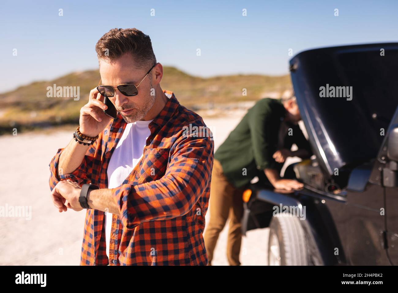Stressed caucasian gay male couple talking on smartphone by broken down car at seaside Stock Photo