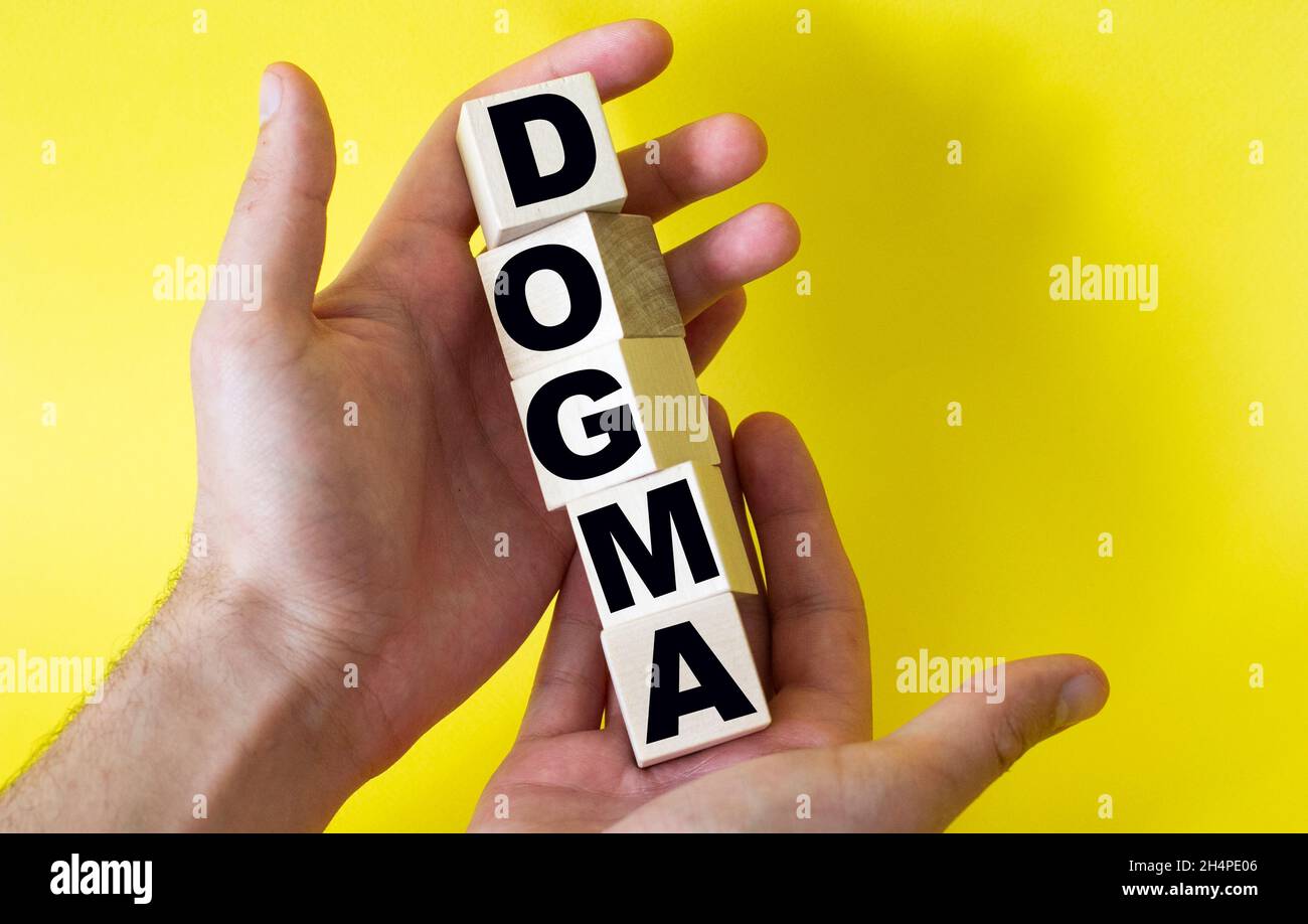 The word DOGMA from building blocks is held by a man on a yellow background Stock Photo