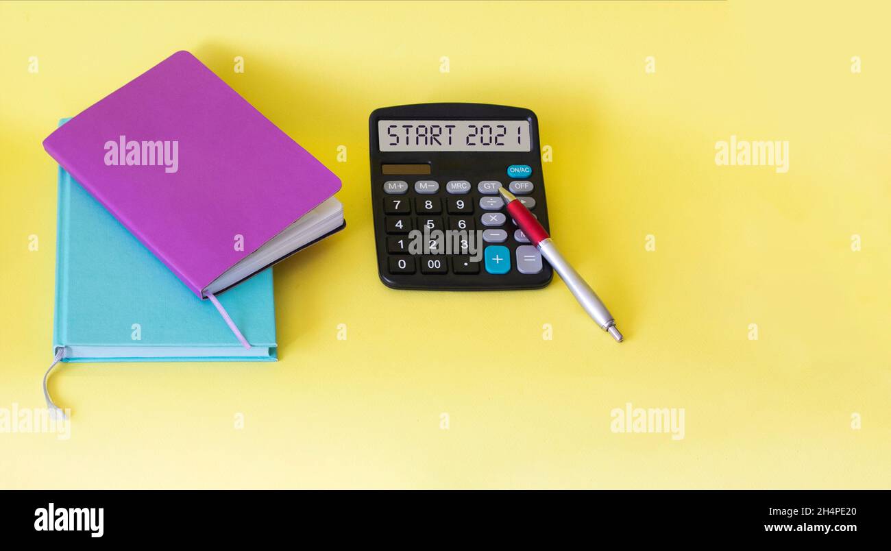 The calculator has the text Start 2021, next to multi-colored notebooks and a pen on a yellow background. Planning, business, motivation and inspirati Stock Photo