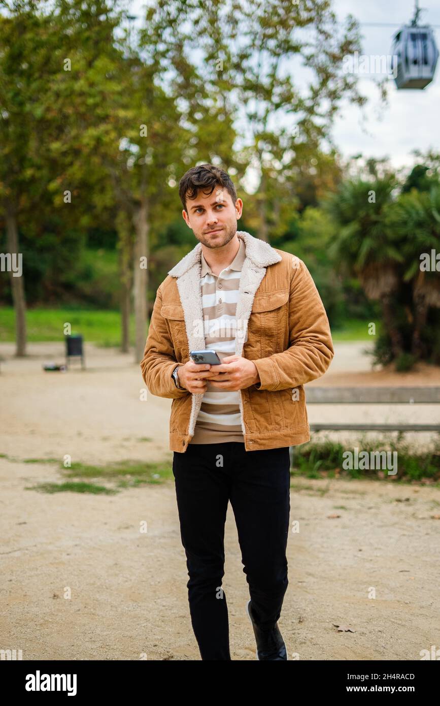 Man in casual outerwear using smartphone and looking away while strolling in park in daytime Stock Photo