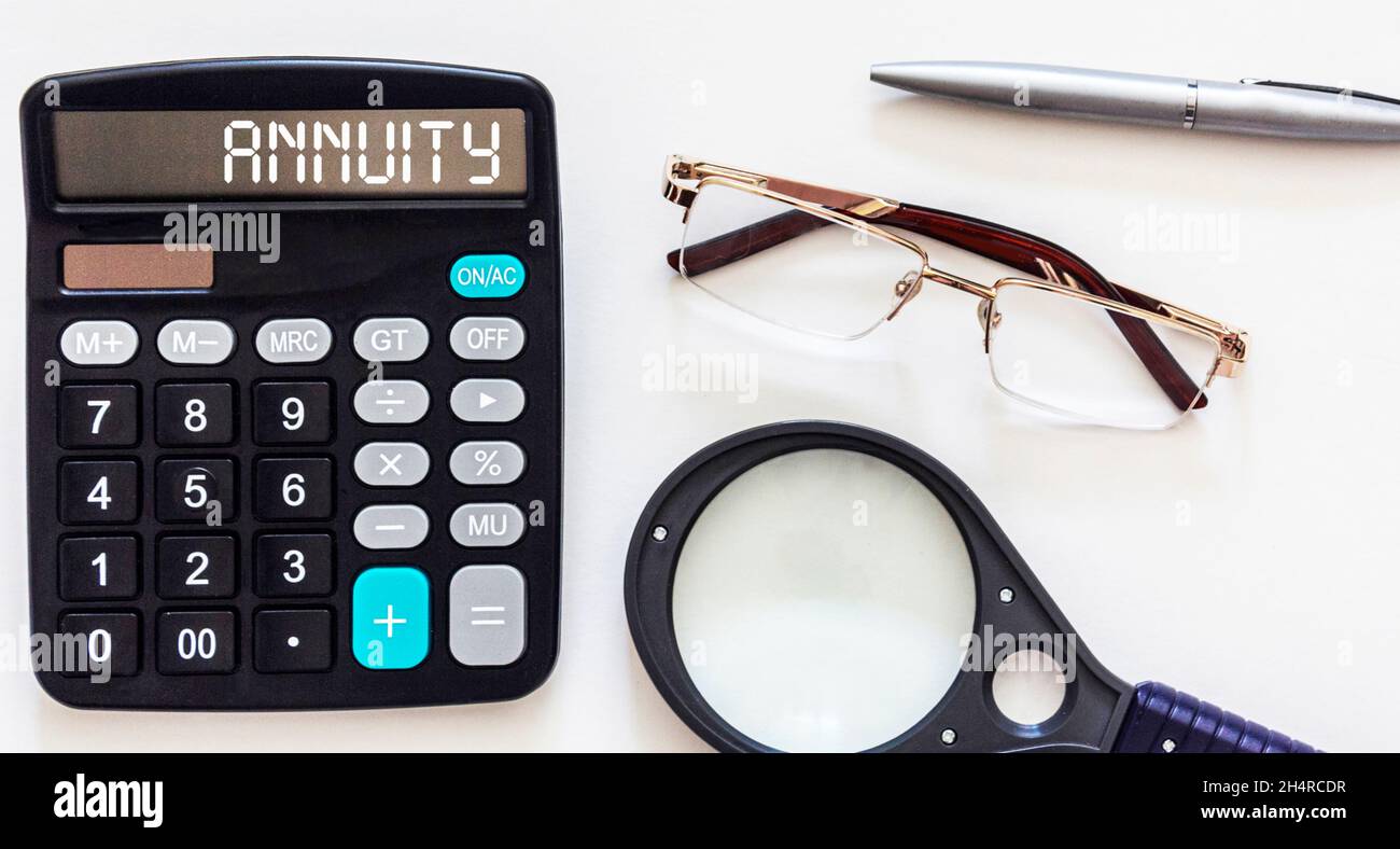 The word ANNUITY is written on a calculator and a white background. Nearby are glasses, a pen, a magnifying glass. Business concept Stock Photo