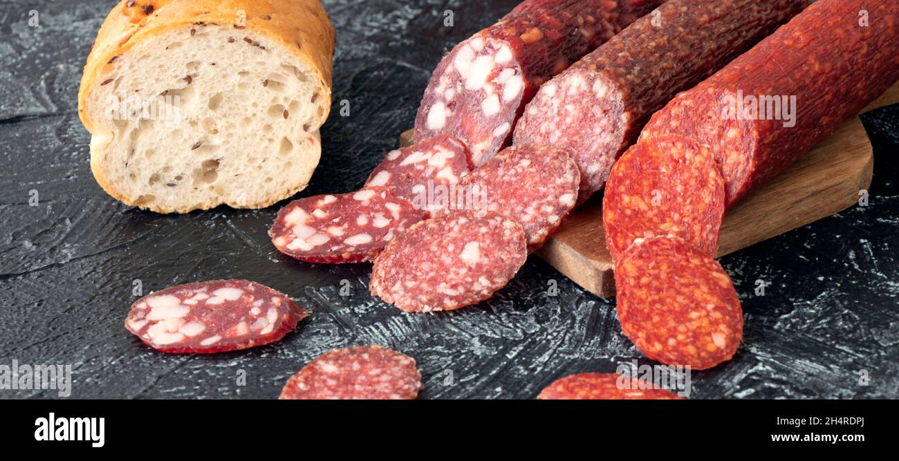 banner with three kind of different sausages on a cutting board near bread. Pieces of sausage cut into circles on a black background. Soft focus. Stock Photo