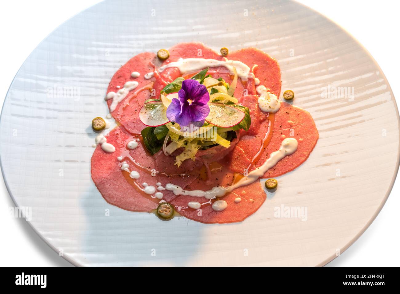 Raw beef carpaccio, Slices of raw veal fassone meat with salad, radishes, capers and violet flower edible in white dish Stock Photo