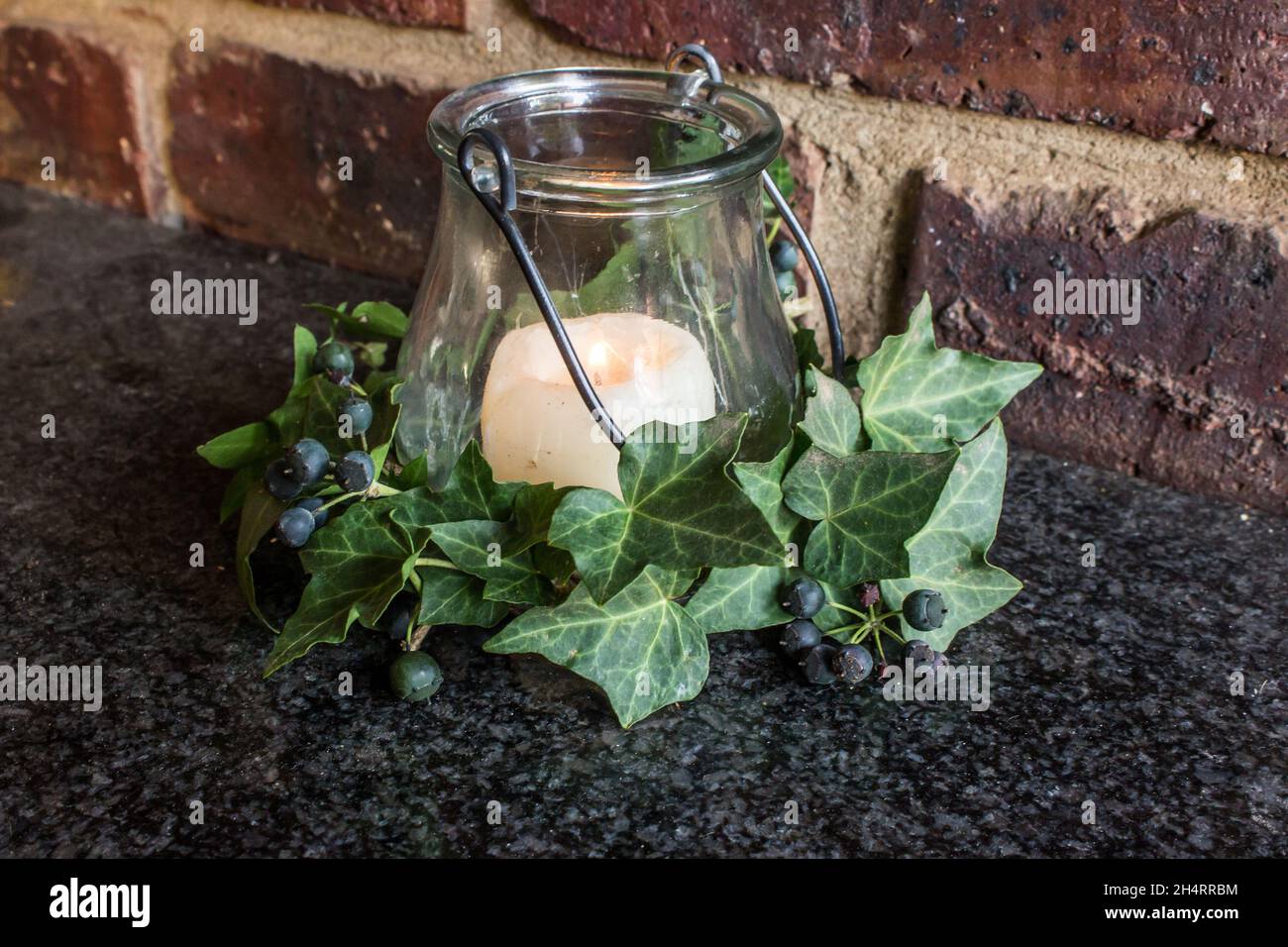 Still-life consisting of a lit candle, surrounded by dark green Ivy leaves and berries, with a rustic brick background. Stock Photo