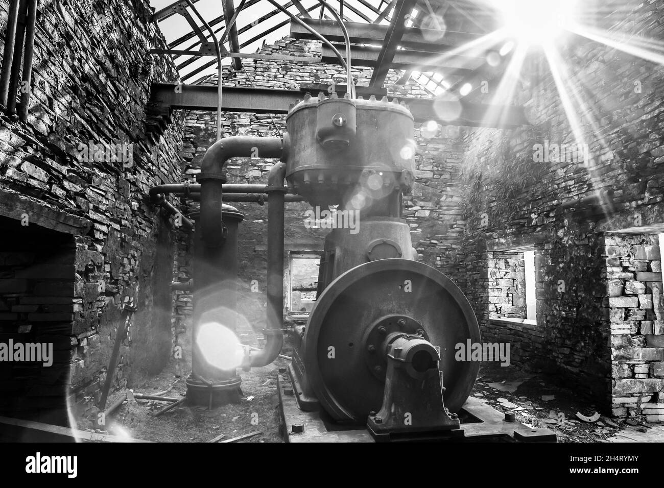 Black and white view of old machinery in derelict, disused slate quarry building, sun streaming through collapsing roof. Stock Photo