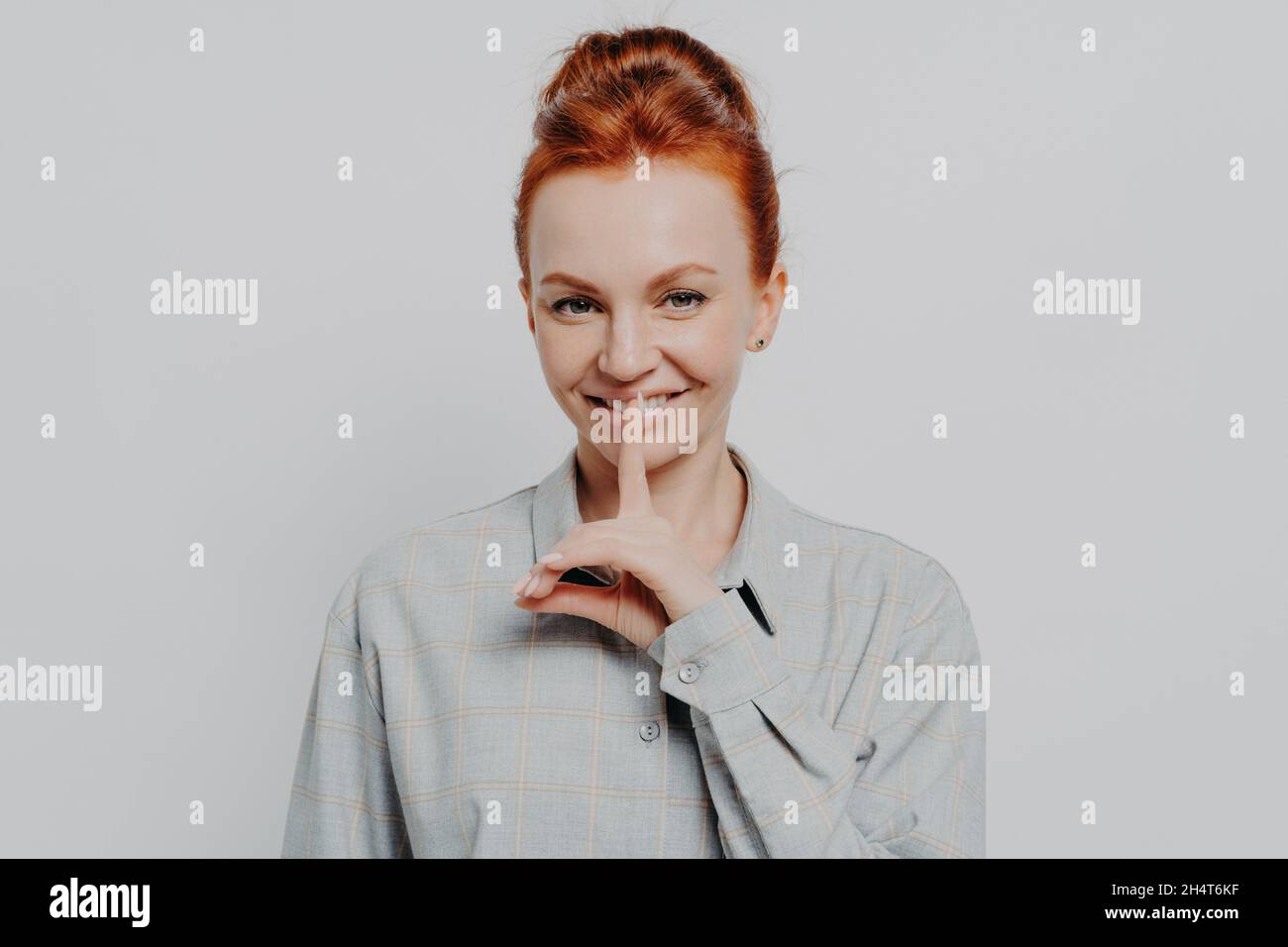 Cheerful young ginger 30s woman showing hush gesture while posing isolated on grey studio background Stock Photo