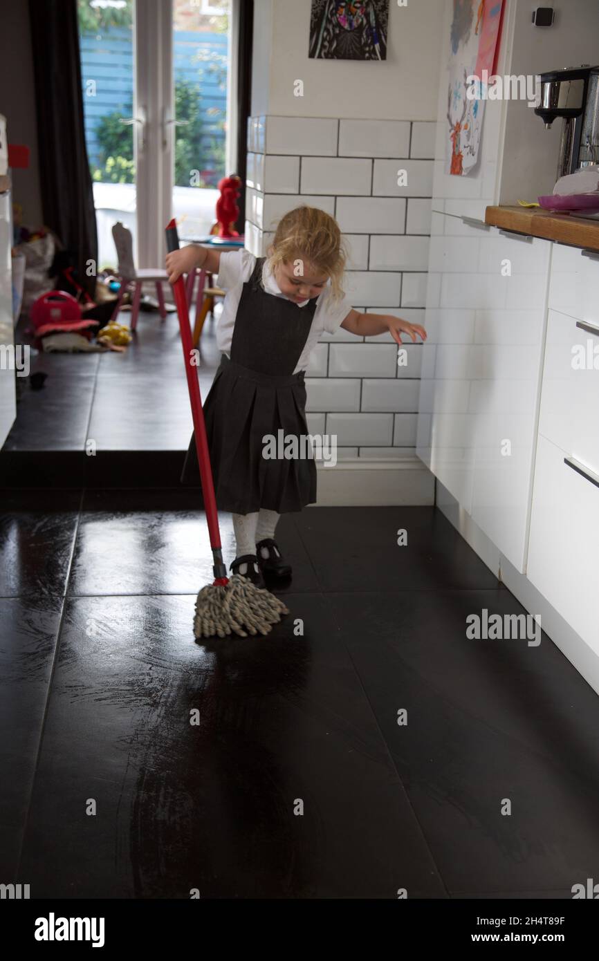A 3 year old girl in school uniform mopping the kitchen floor, UK Stock Photo