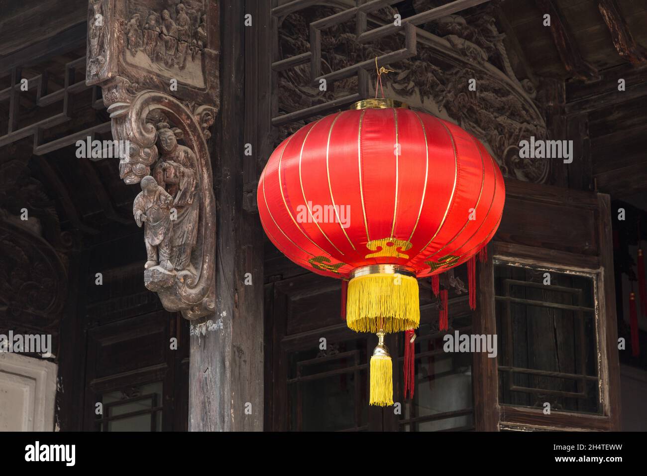 A red Chinese silk lantern hangs by an old wood carving on a historic building in Wuzhen, China. Stock Photo