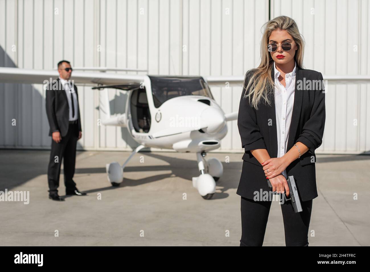 Beautiful professional female spy agent bodyguard with gun securing airplane on private airport. Security police woman in civilian black suit. Close s Stock Photo