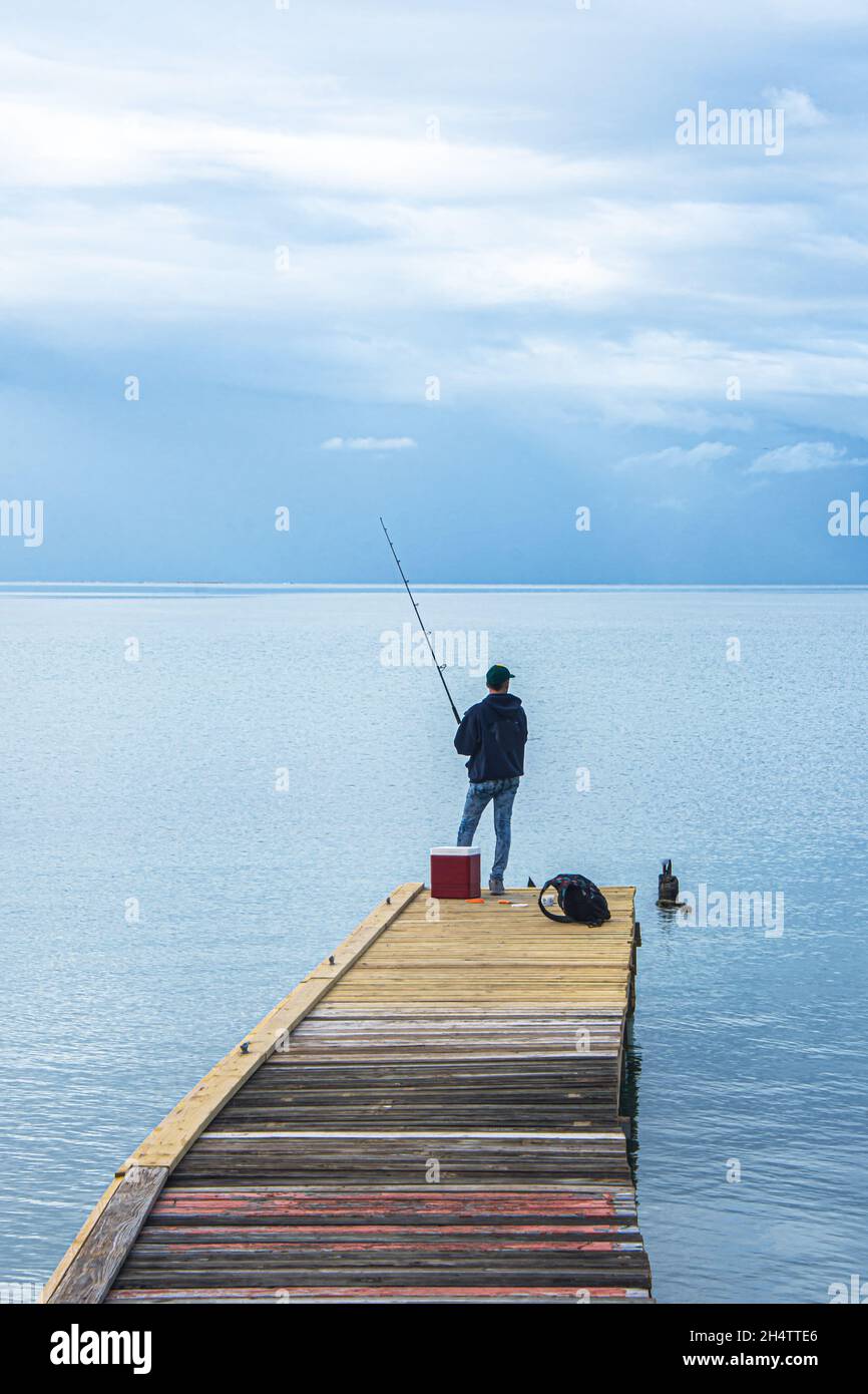 Young man fishing on dock with Caribbean Sea stretching to grey sky horizon. Vertical format. Salinas, Puerto Rico, USA. Stock Photo