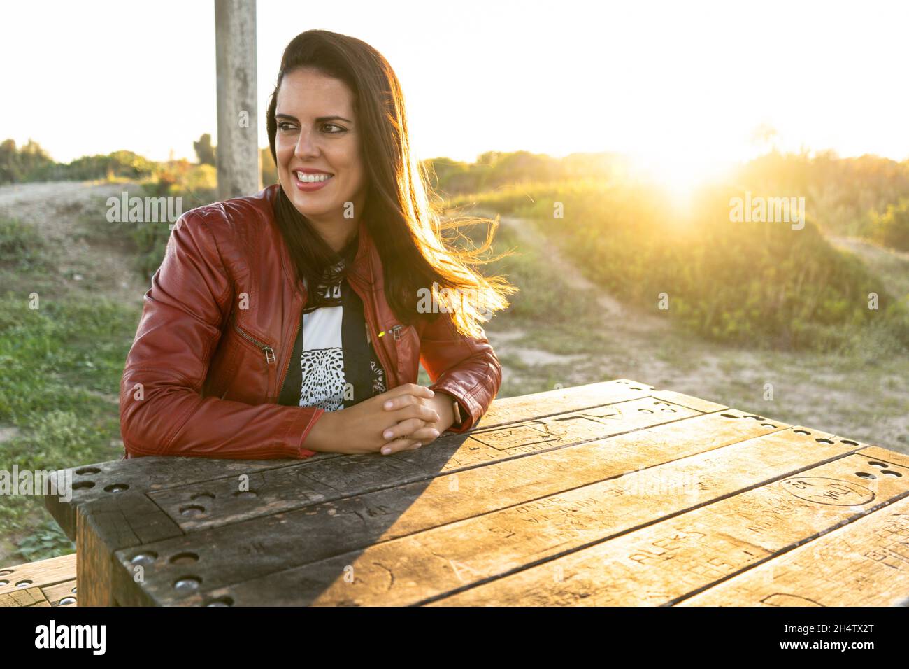 Young woman smiling sitting at a table in a park at sunset wearing a red jacket Stock Photo