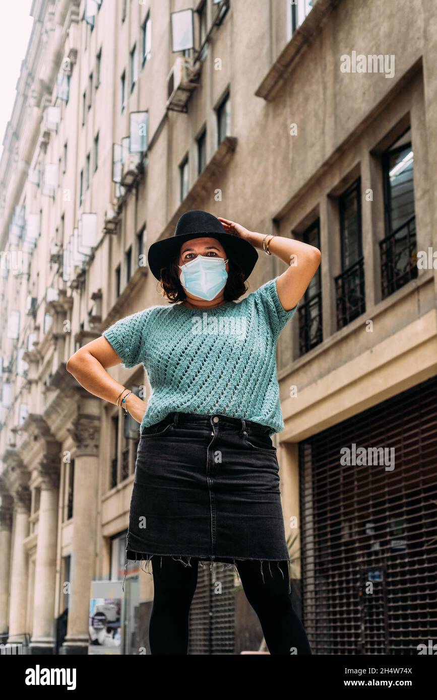 Woman wearing a hat and protecting herself with a blue mask, she is fashionable and has buildings of ancient architecture in the background. Covid 19 Stock Photo