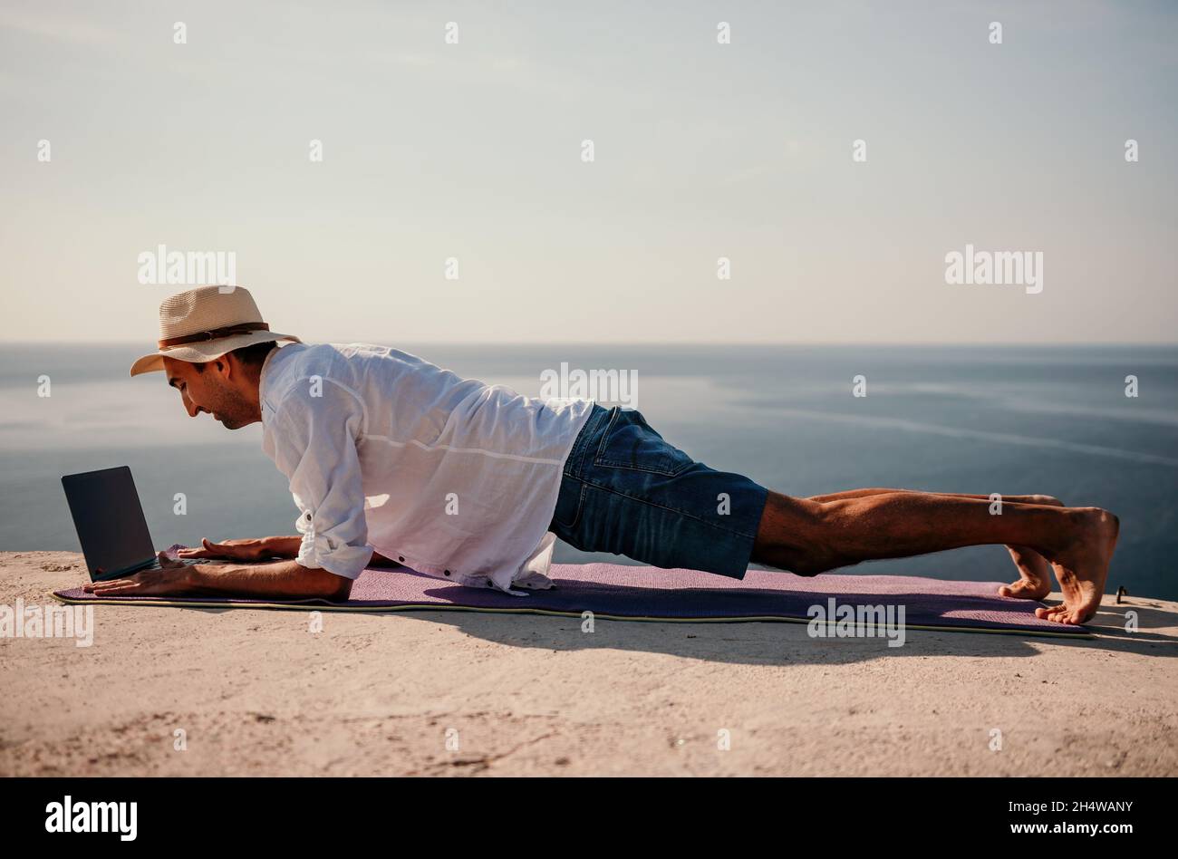 A digital nomad, a man in a hat, a businessman with a laptop does yoga on the rocks by the sea at sunset time, does a business operation online from a Stock Photo