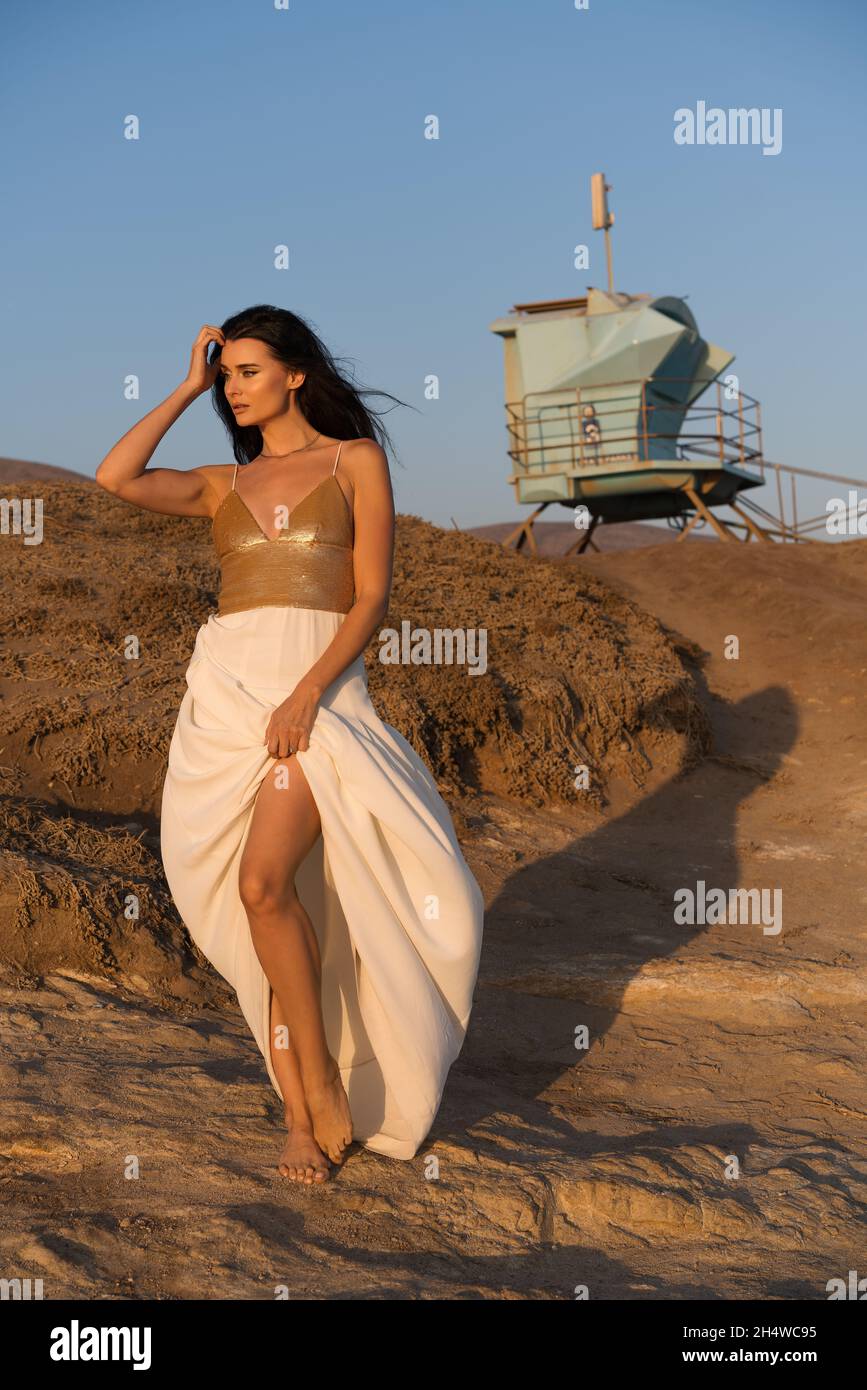 A beautiful and sexy black hair female modeling wearing a gold white evening gown, posing and standing on a rocky cliff at sunset. Stock Photo