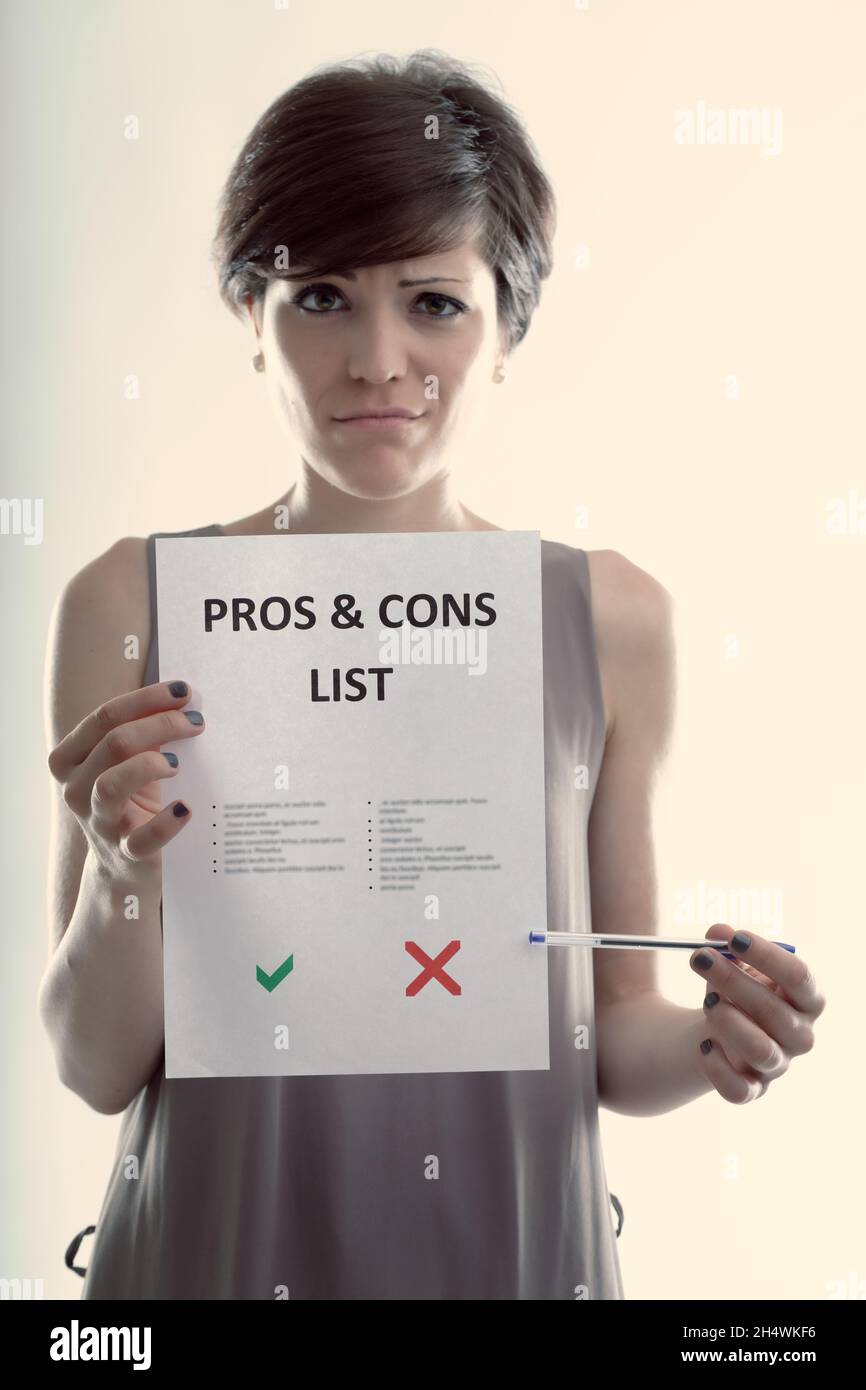 Cute young woman holding up a Pros and Cons list with a wry smile pointing to the cons which has more points against Stock Photo