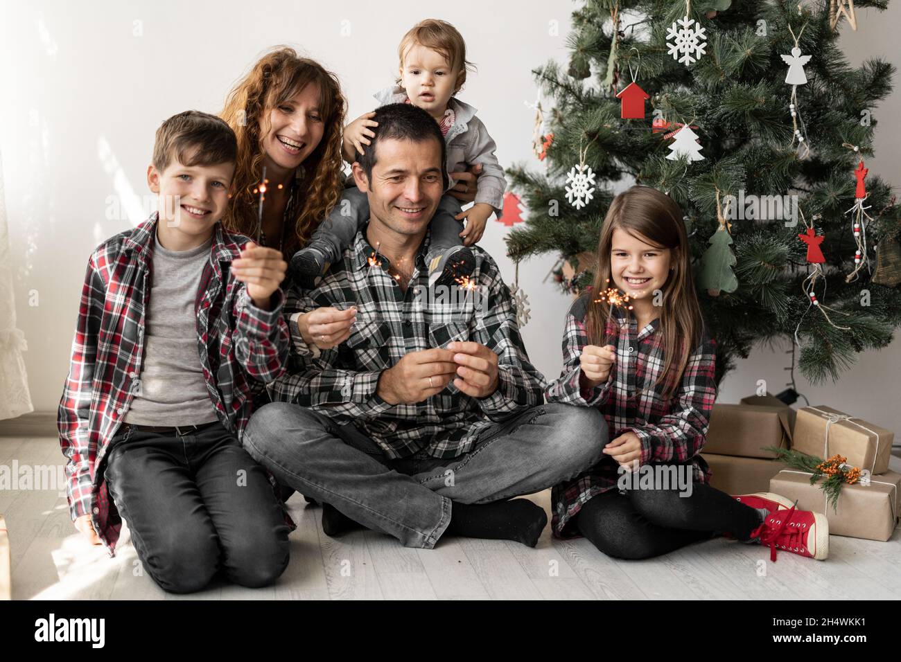 Happy full family and three children at home with Christmas gifts sitting at home on the floor in the morning under a Christmas tree in winter. Stock Photo