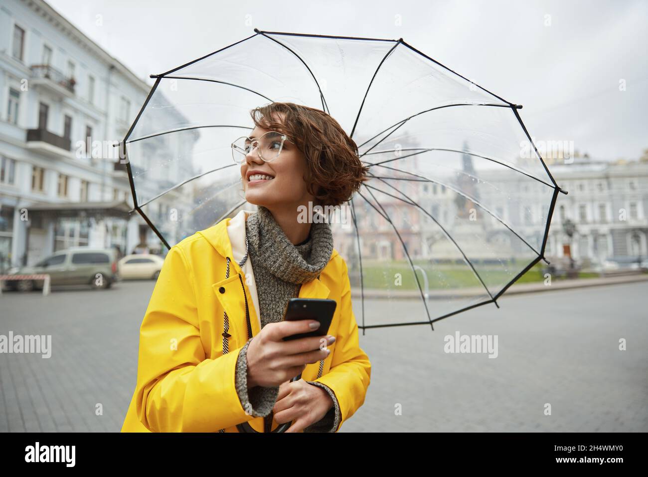 Caucasian girl in yellow raincoat with transparent umbrella in city. Young beautiful woman
