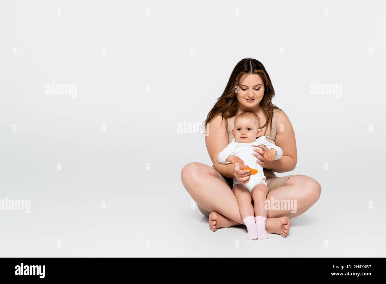 Cheerful body positive woman hugging baby daughter while sitting on grey background Stock Photo