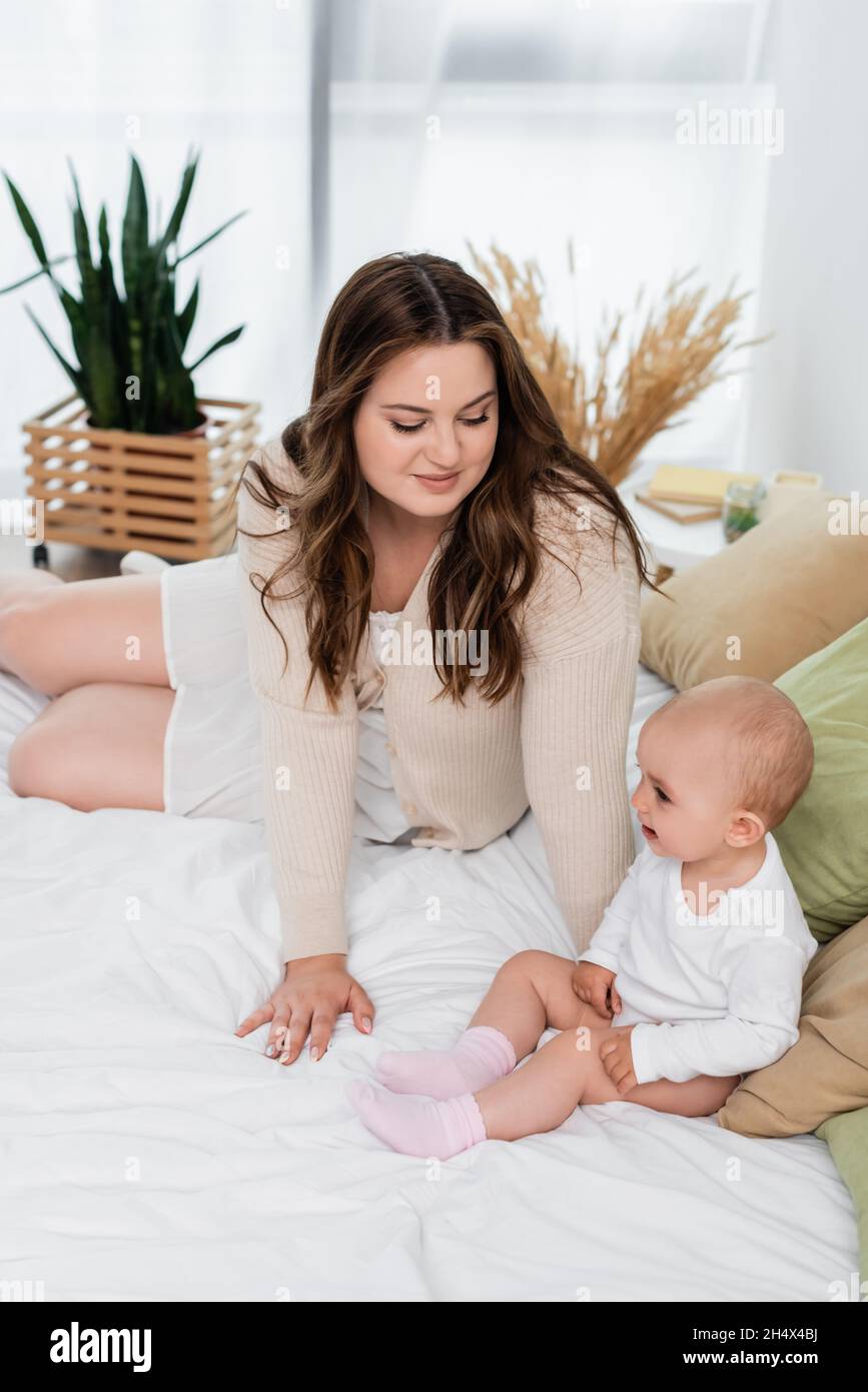 Young body positive mother looking at cheerful baby on bed Stock Photo