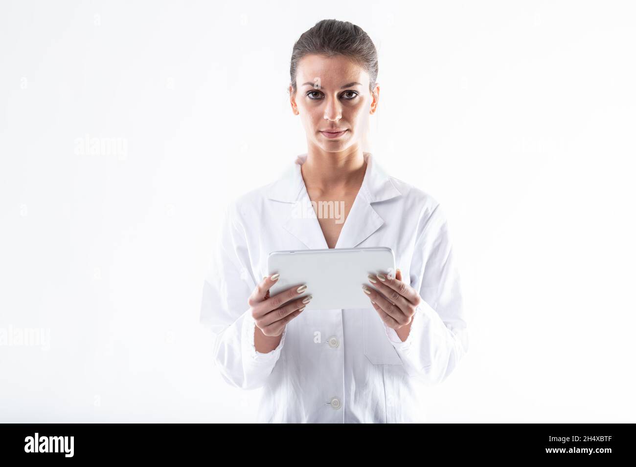 Attractive young female doctor or nurse in a white lab coat standing looking at the camera holding a tablet-pc with a serious thoughtful expression is Stock Photo