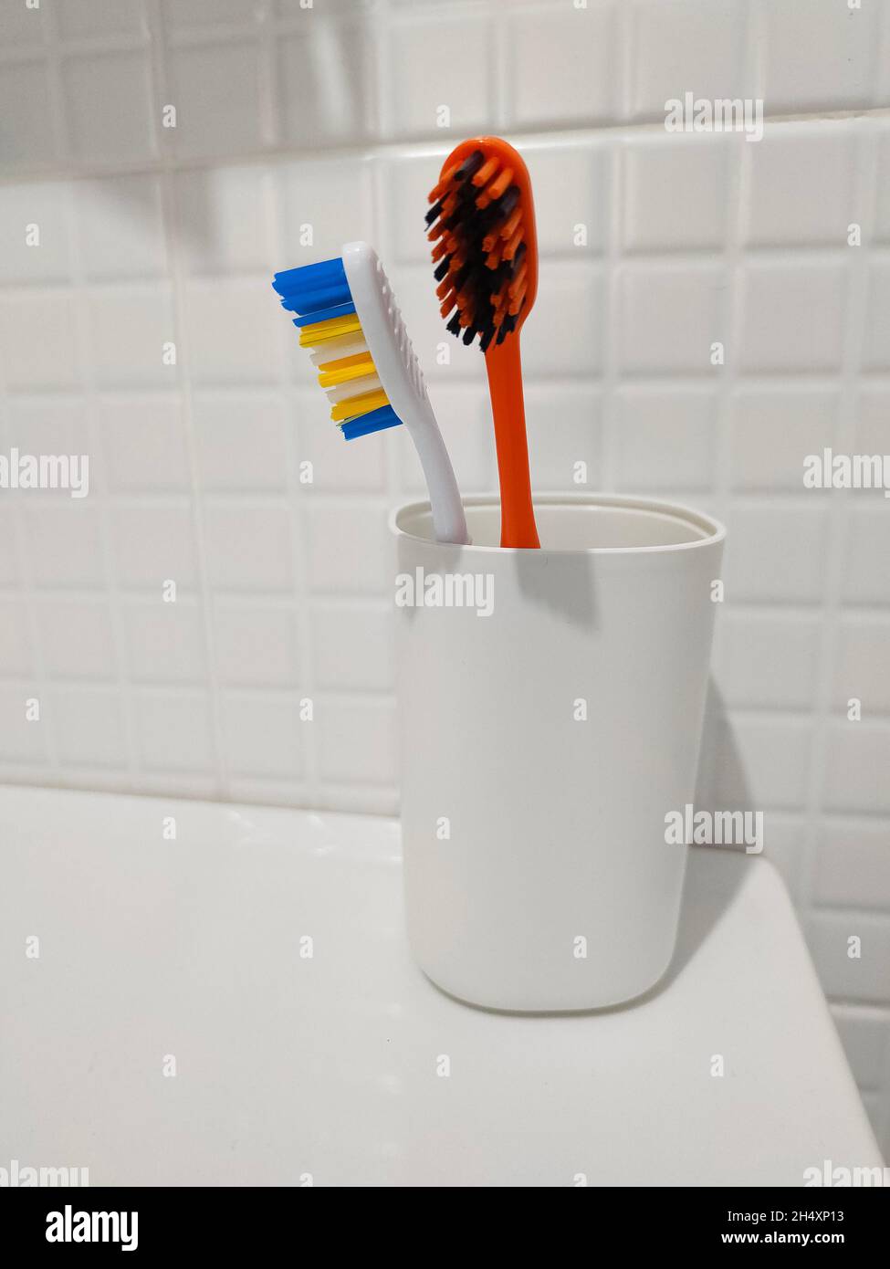 Two colored toothbrushes in white plastic glass on the shelf. Stock Photo
