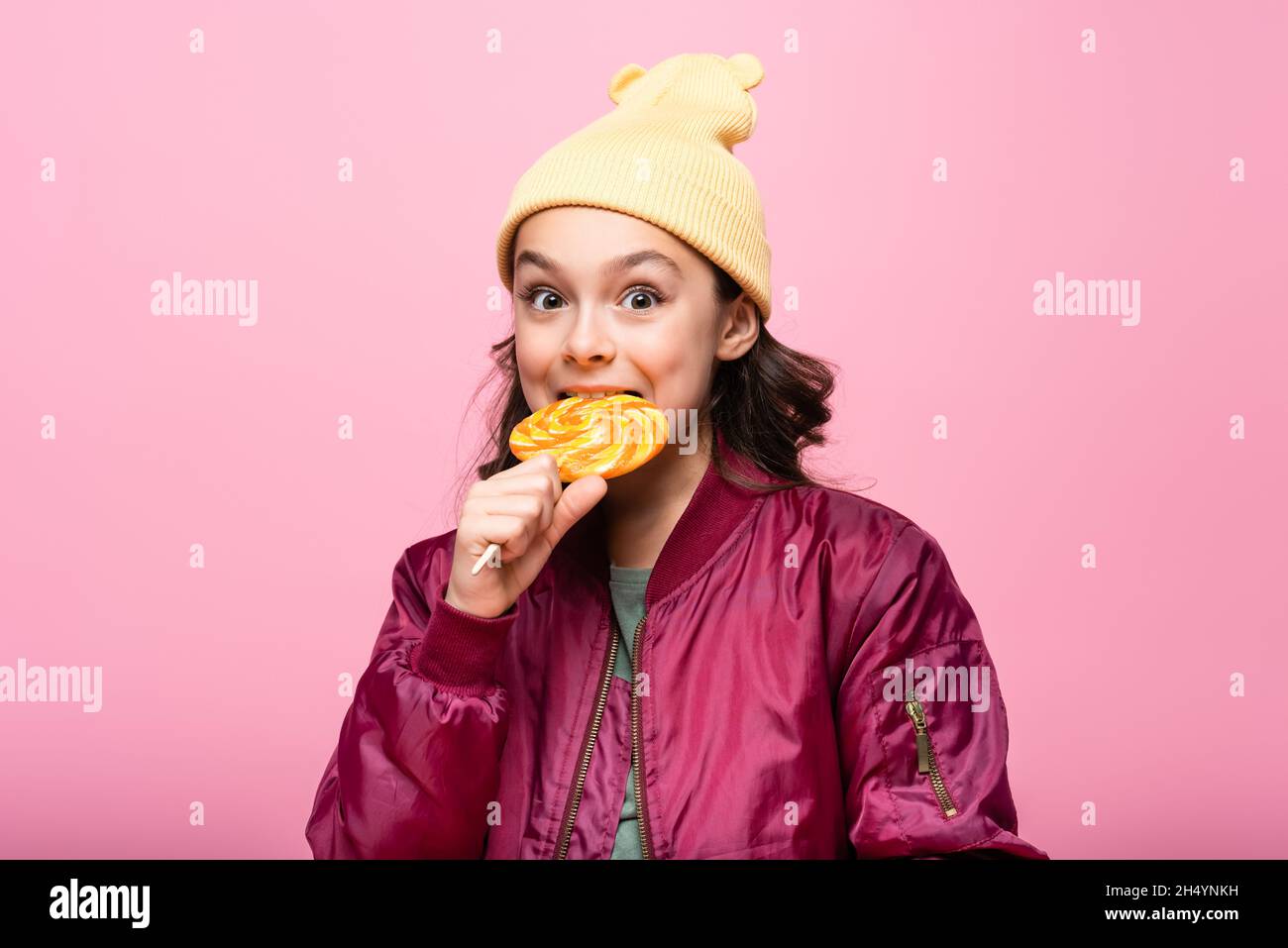 trendy preteen girl in winter outfit biting lollipop isolated on pink Stock Photo