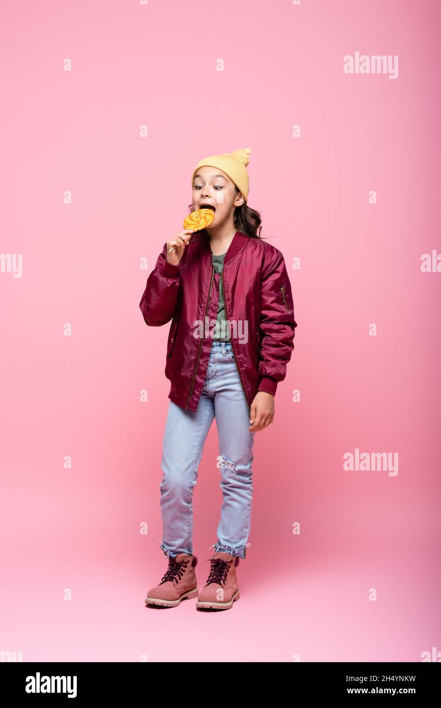 full length of preteen girl in stylish outfit eating lollipop on pink Stock Photo