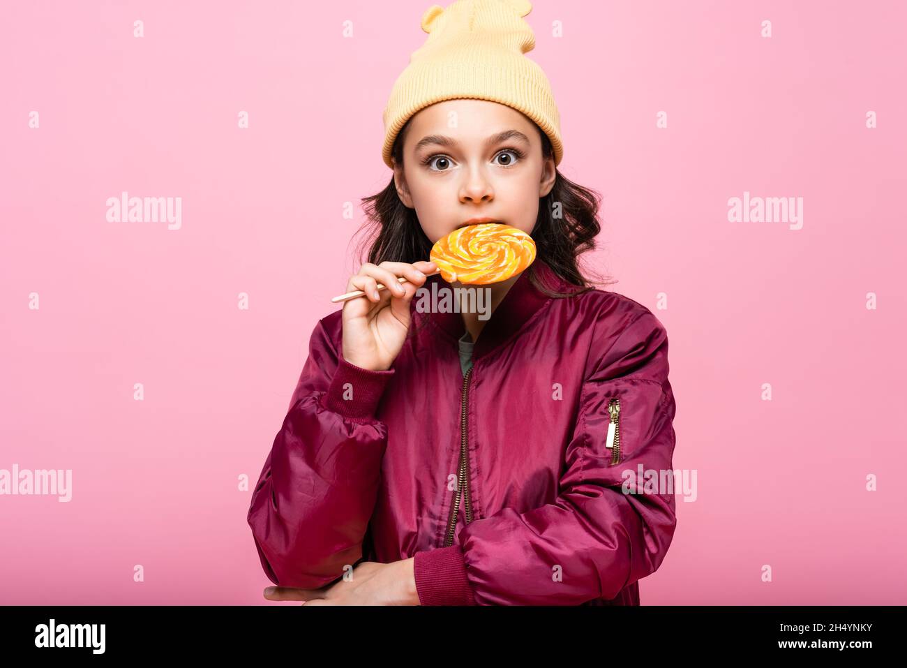 stylish preteen girl in winter outfit biting lollipop isolated on pink Stock Photo