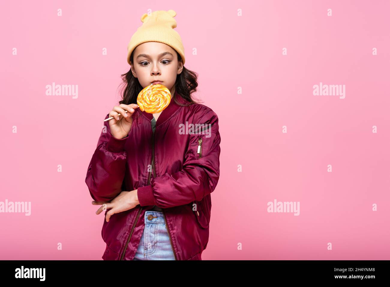 stylish preteen girl in winter outfit looking at lollipop isolated on pink Stock Photo