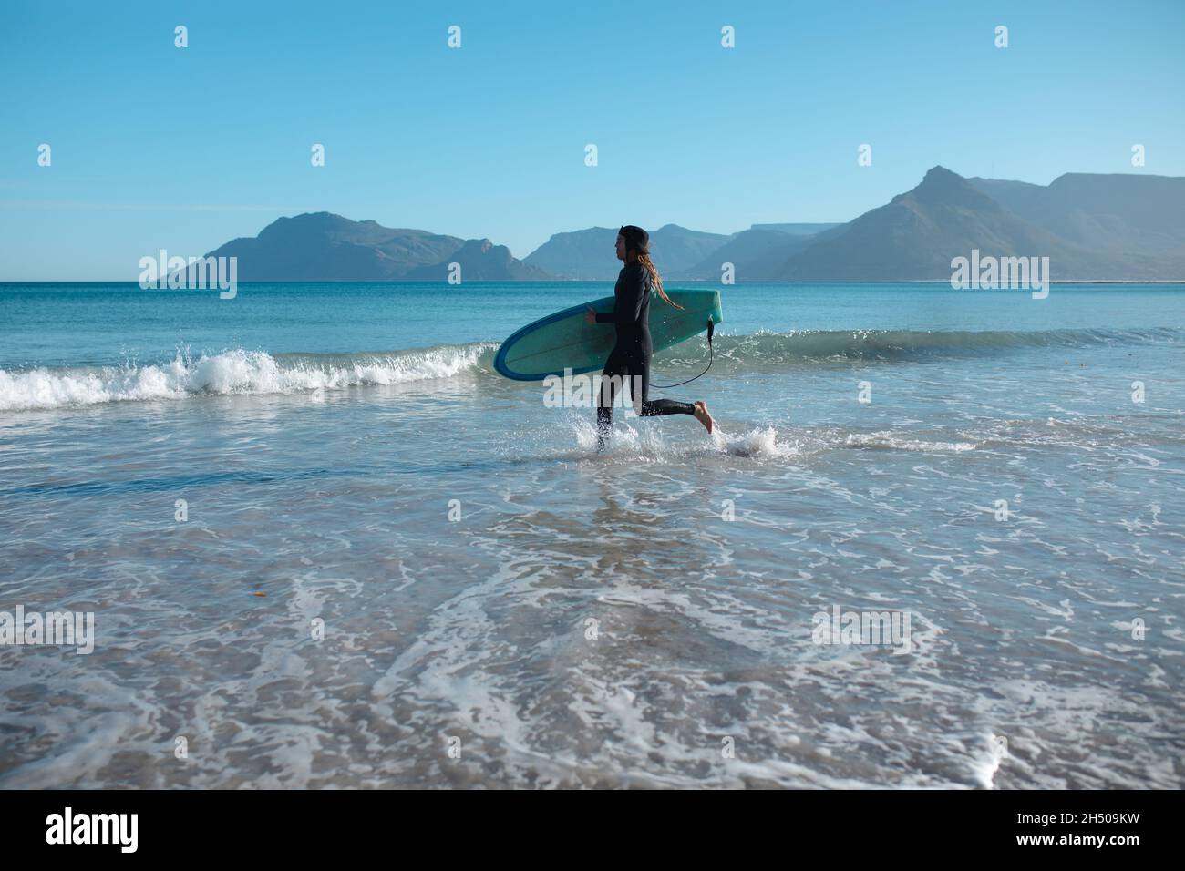 Man carrying surfboard running while splashing water on shore against blue sky with copy space Stock Photo