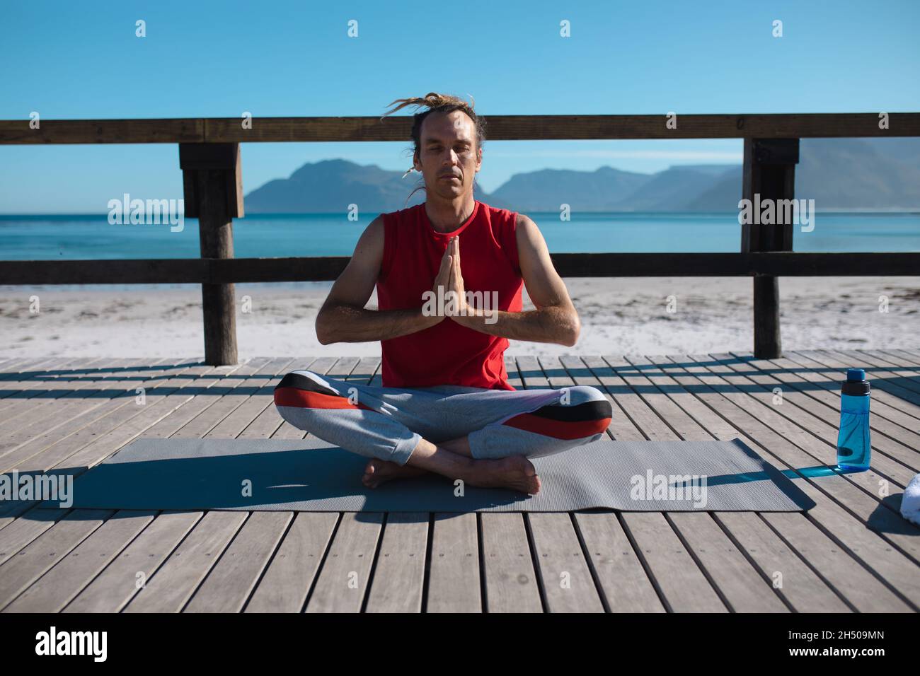 Man practicing yoga while sitting cross-legged and hands clasped meditating on floorboard at beach Stock Photo