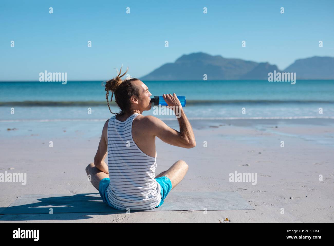 Rear view of man drinking water from bottle while doing yoga at beach on sunny day Stock Photo