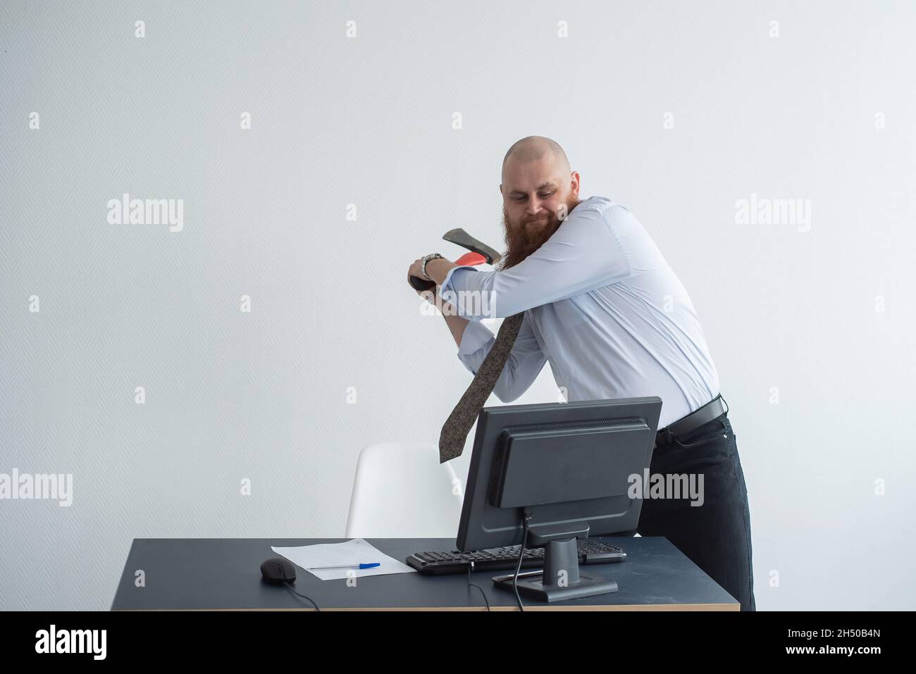Angry bald man with a red beard in the office in a business suit smashes an ax with a computer. The manager with a nervous breakdown, breaks the Stock Photo