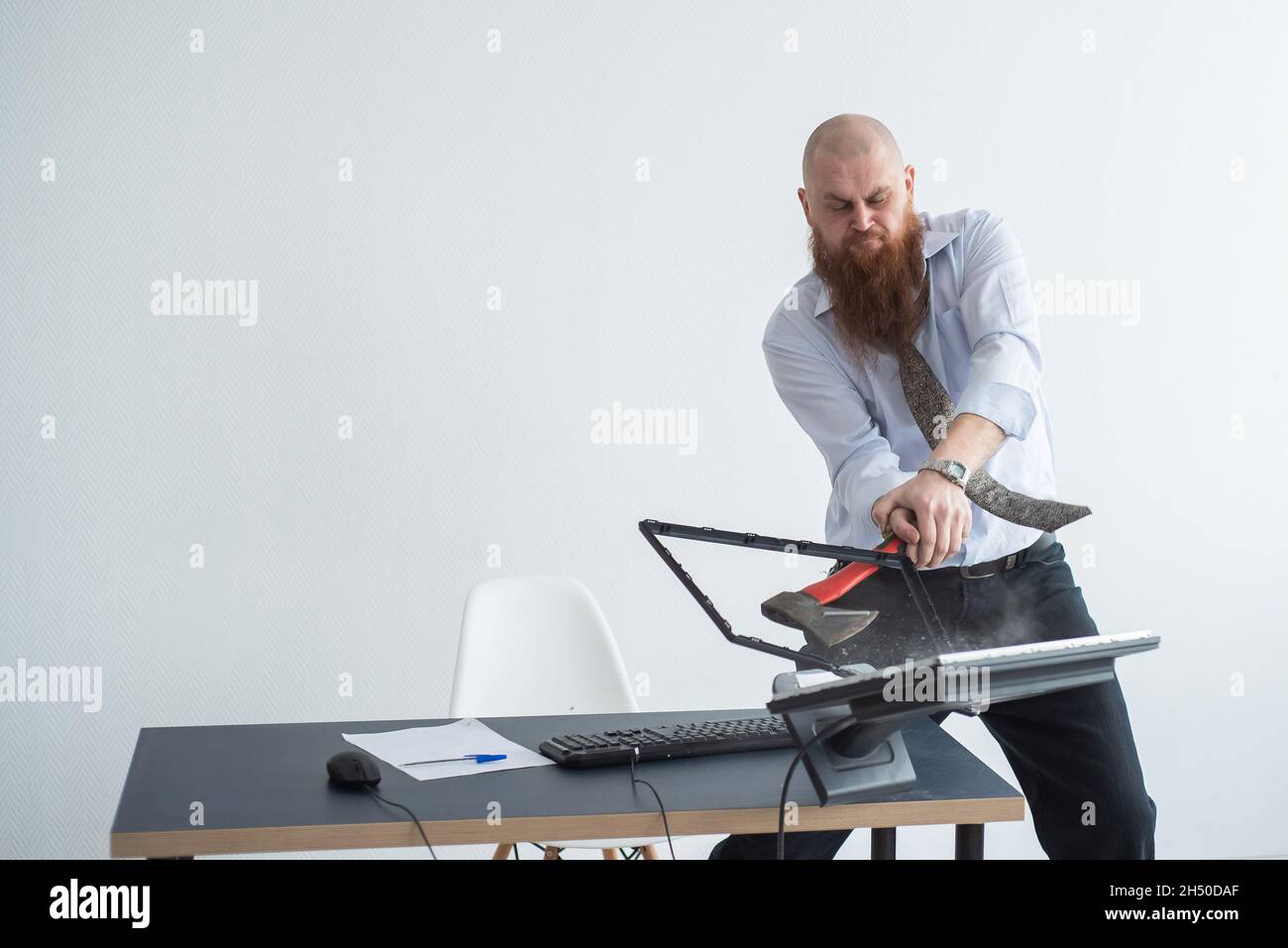 Angry bald man with a red beard in the office in a business suit smashes an ax with a computer. The manager with a nervous breakdown, breaks the Stock Photo