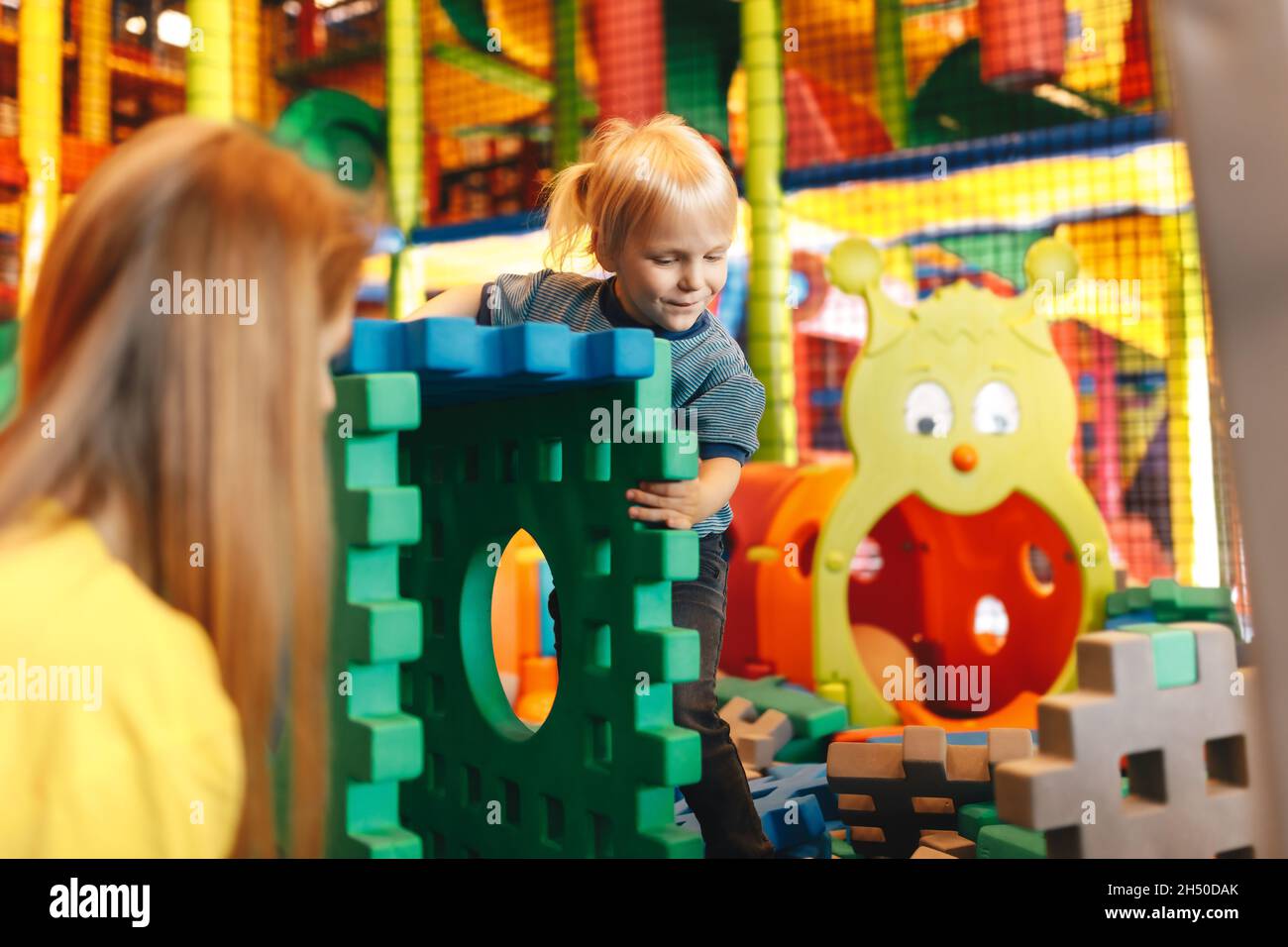 Happy Kid Playing With Big Puzzles in a Playground Center. Child With Young Mother Having Fun Together. Boy Contructing House With Toy Blocks Stock Photo