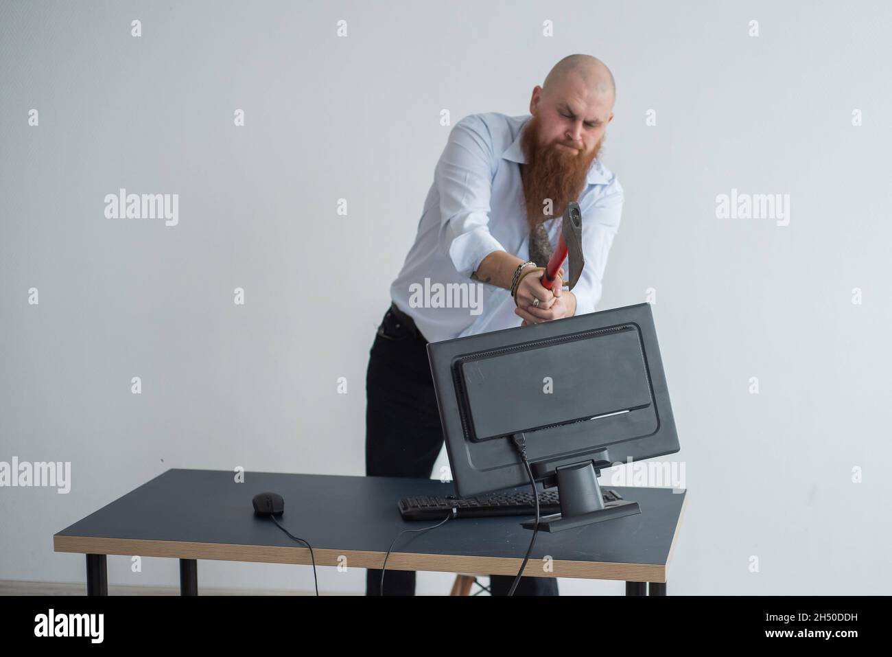 Angry bald man with a red beard in the office in a business suit smashes an ax with a computer. The manager, focused with a nervous breakdown, breaks Stock Photo