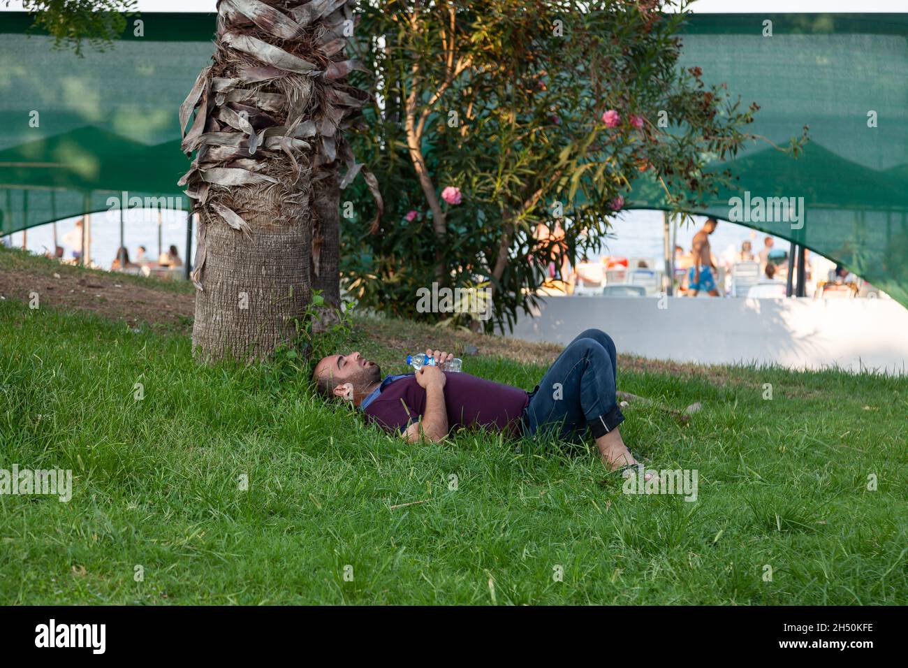 Kemer, Turkey - 08. 25. 2021: A man who fell asleep on a green lawn in the summer sun. A homeless man sleeps on the grass in the park. Stock Photo