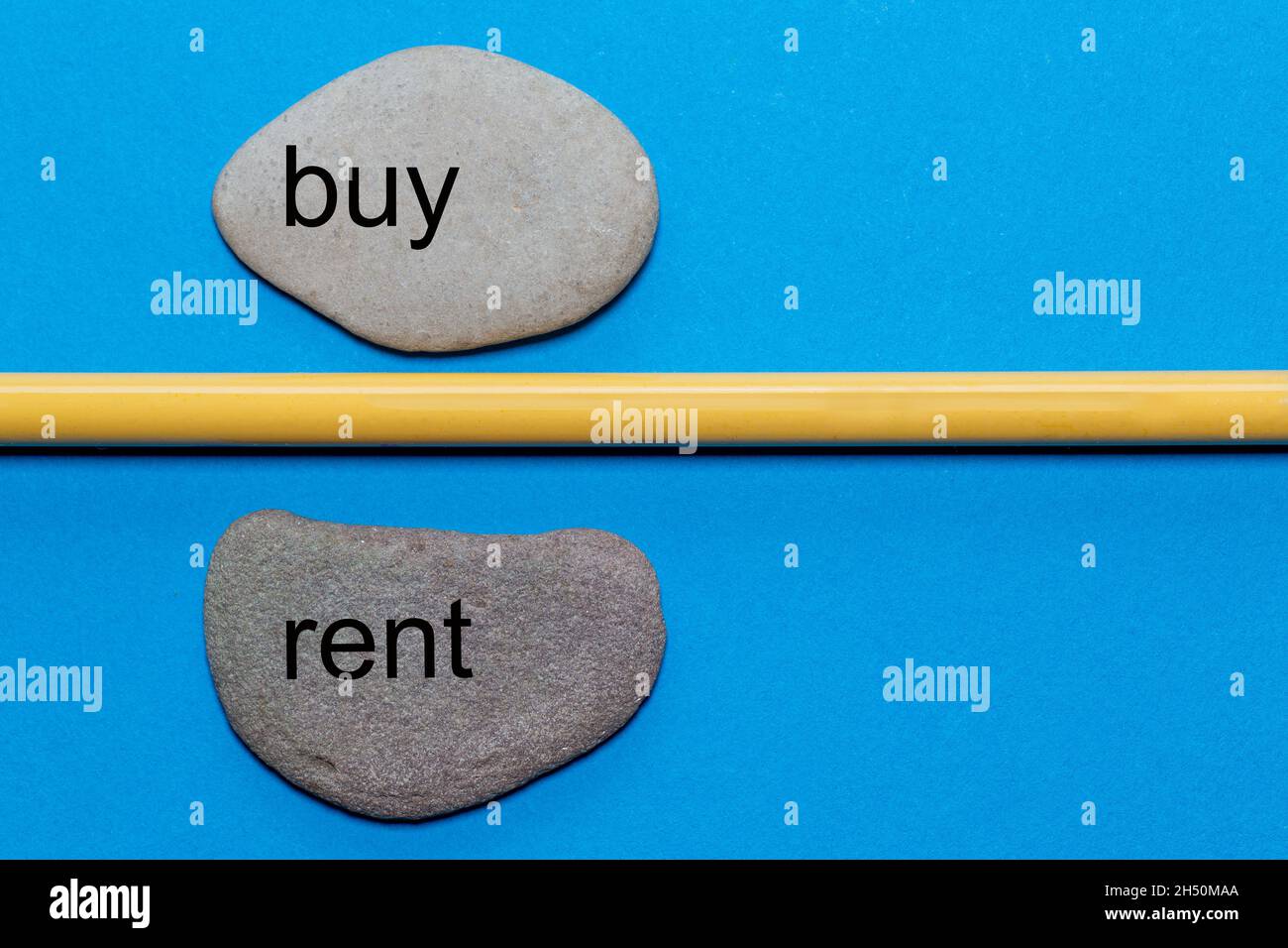 The words buy and rent are written on natural smooth stones separated by a yellow pencil. The background is isolated in blue and has a lot of space Stock Photo