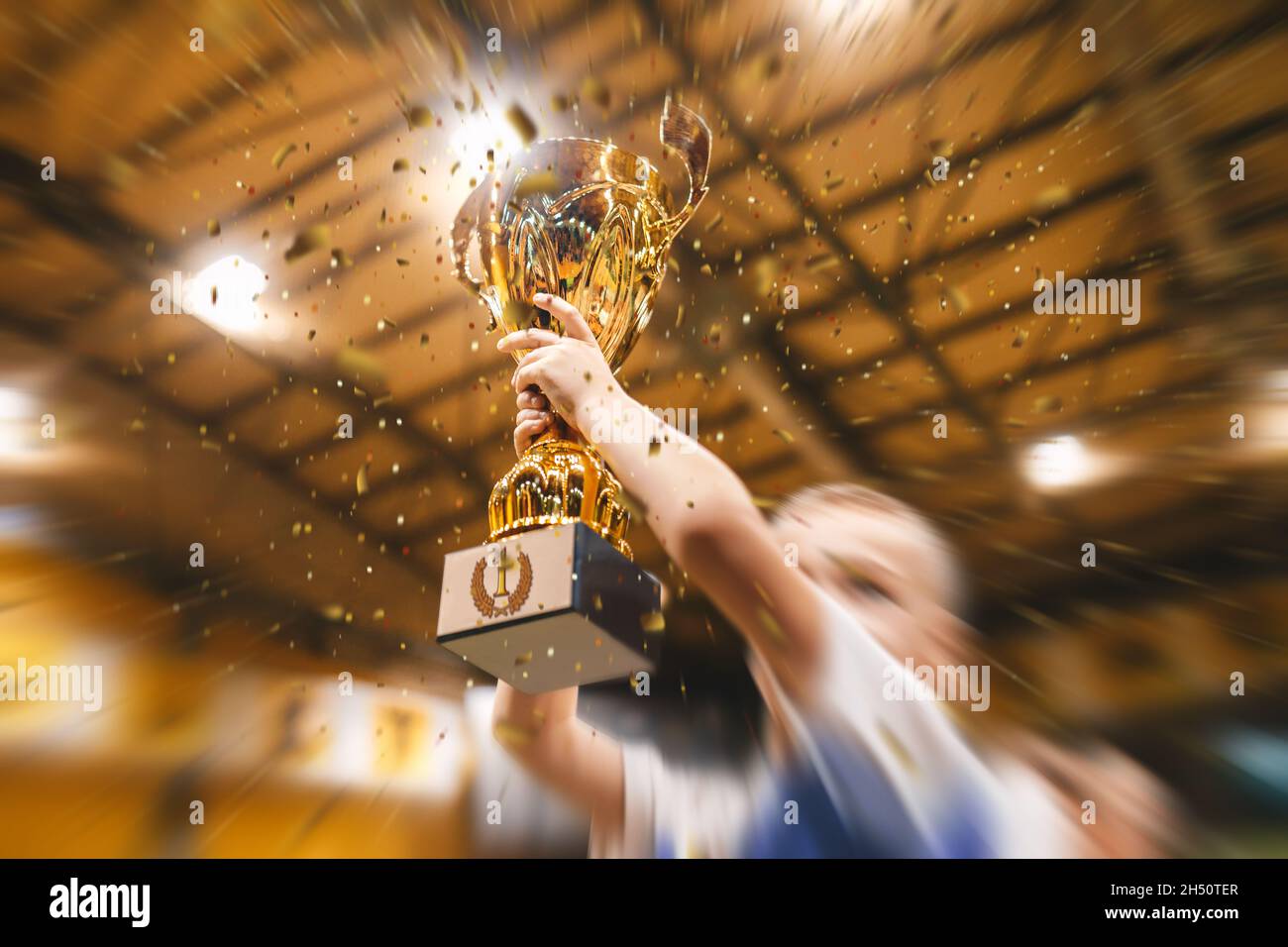 Happy boy rising golden cup, indoor sports celebration championship. Golden confetti falling. Kids in the sports team lift up the golden cup trophy af Stock Photo