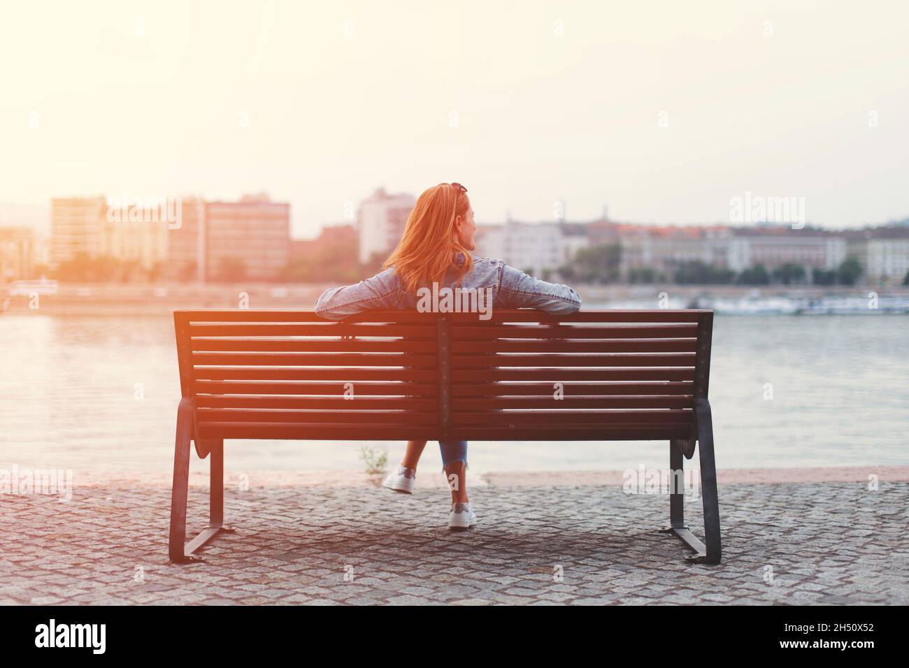 Young redhead woman relaxing on bench at riverside in sunset Stock Photo